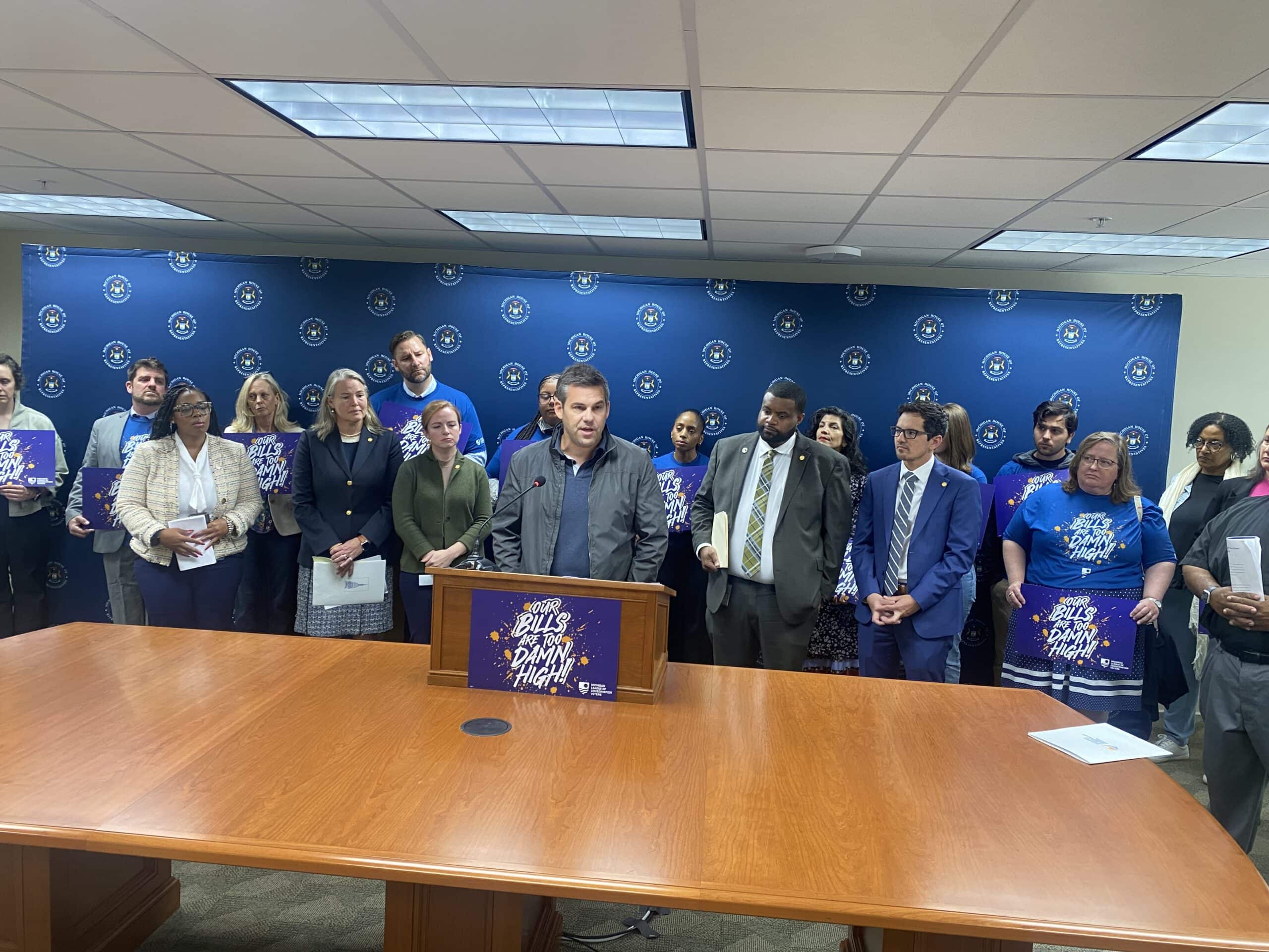 Michigan League of Conservation Voters' Deputy Director Bob Allison stands at a podium with a sign that says "Our Bills Are Too Damn High" with a group of Michigan lawmakers and community leaders standing behind him.